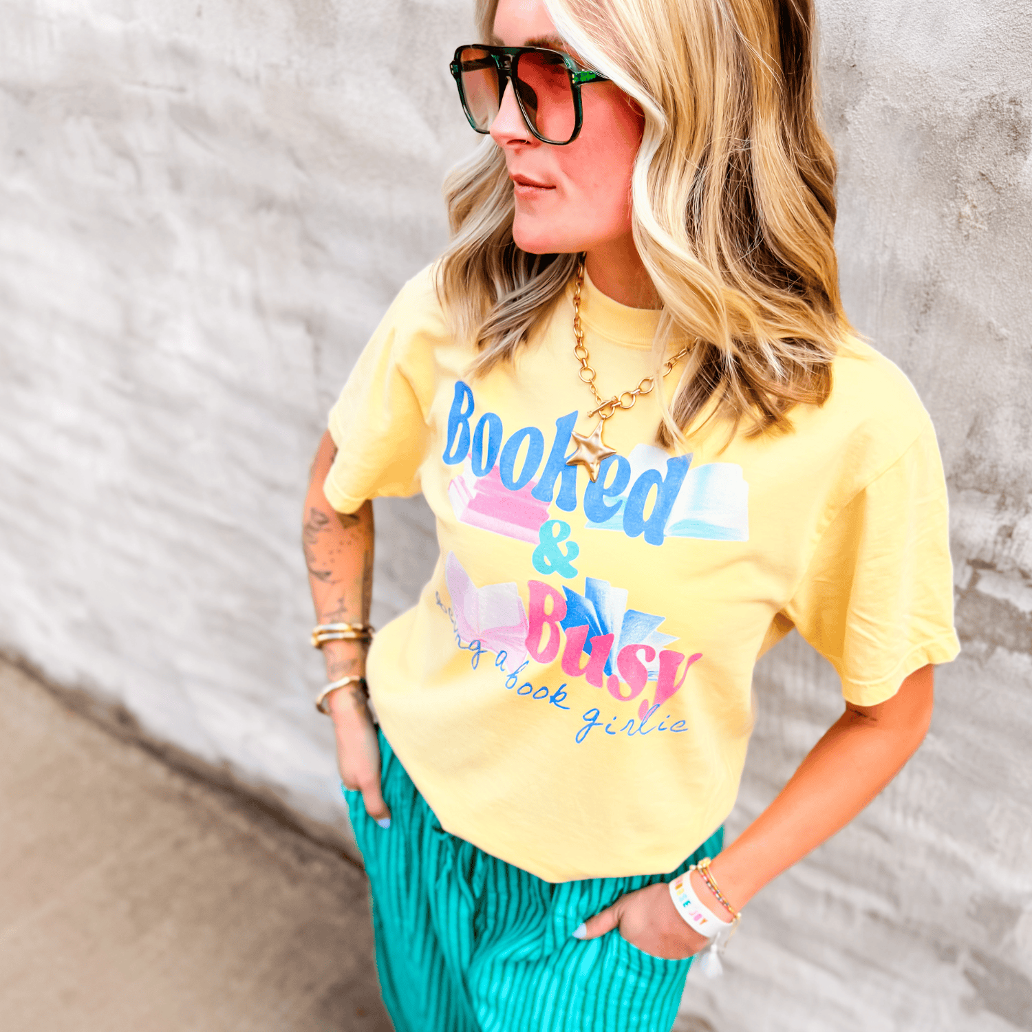 Yellow "Booked & Busy" tee with colorful text, worn by a woman with wavy hair, paired with teal striped pants.