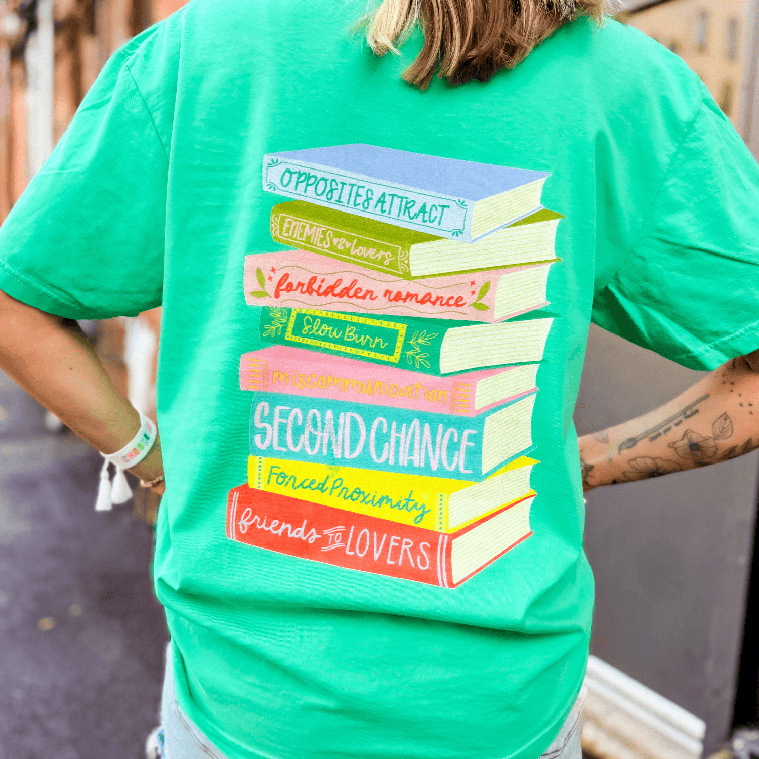 Just Here For The Tropes Tee in bright green featuring colorful book stack graphic with romance tropes on the back.