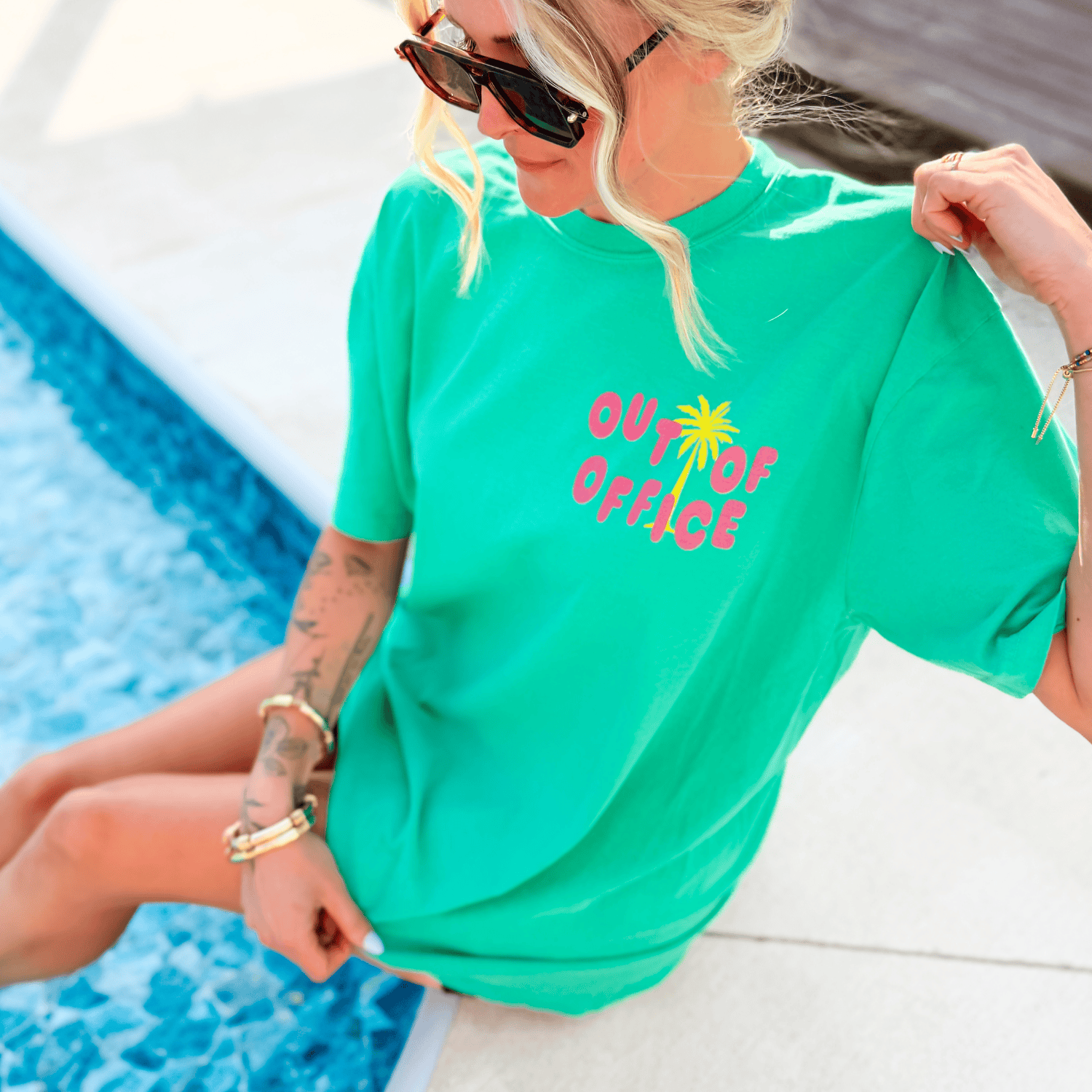 Neon green tee featuring "Out of Office" text with a palm tree design, worn by a person sitting by a pool.