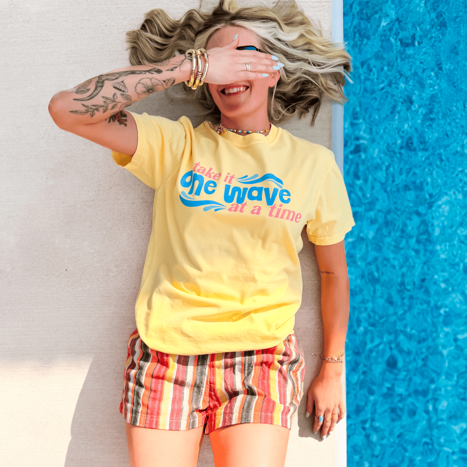 Yellow One Wave Tee with a colorful graphic, worn by a smiling person lying beside a blue pool.