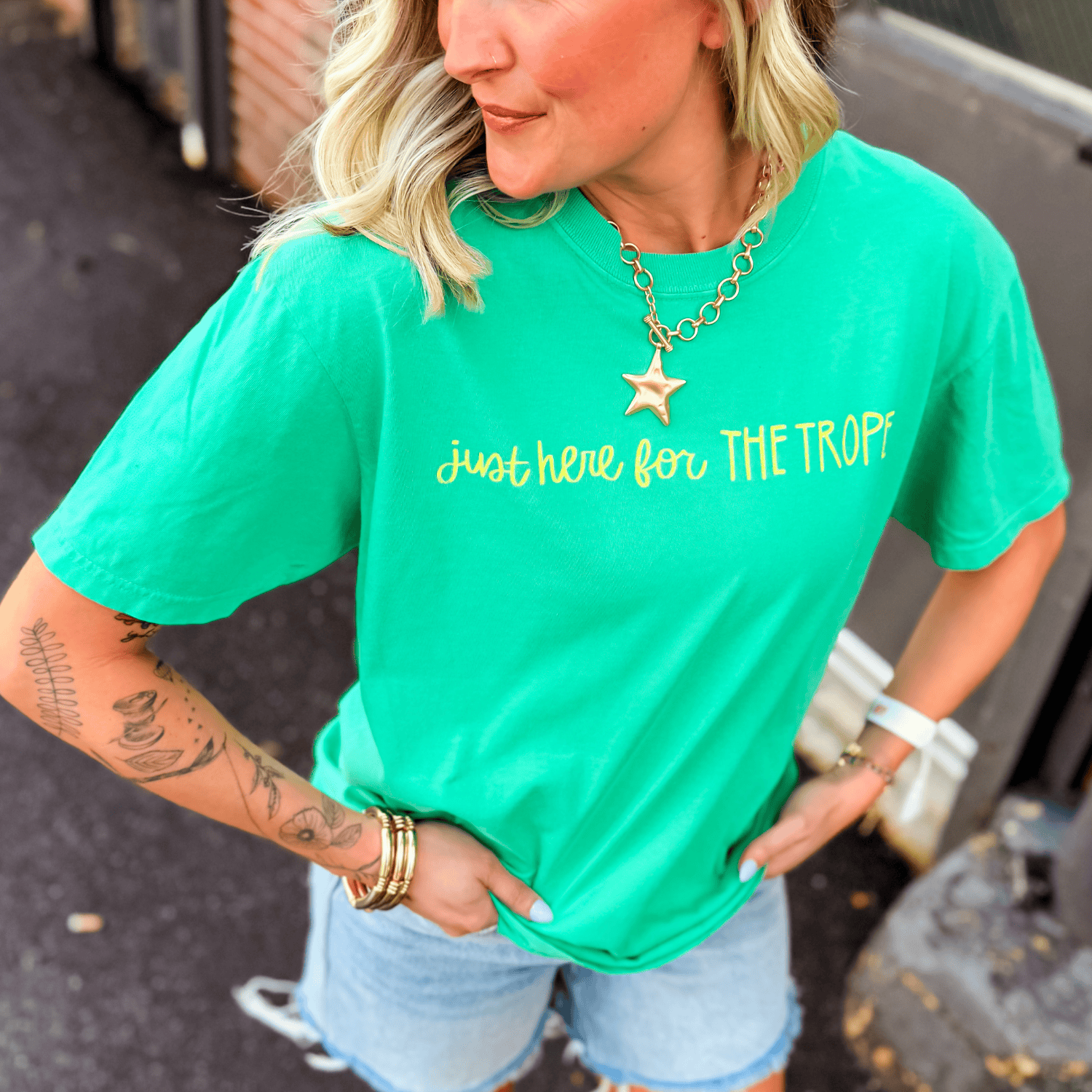Green t-shirt with "just here for THE TROPES" in yellow text, styled with denim shorts and accessories.