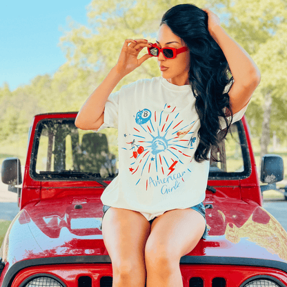 American Girlz tee featuring a colorful graphic design, worn by a woman with long hair, sitting on a red jeep.