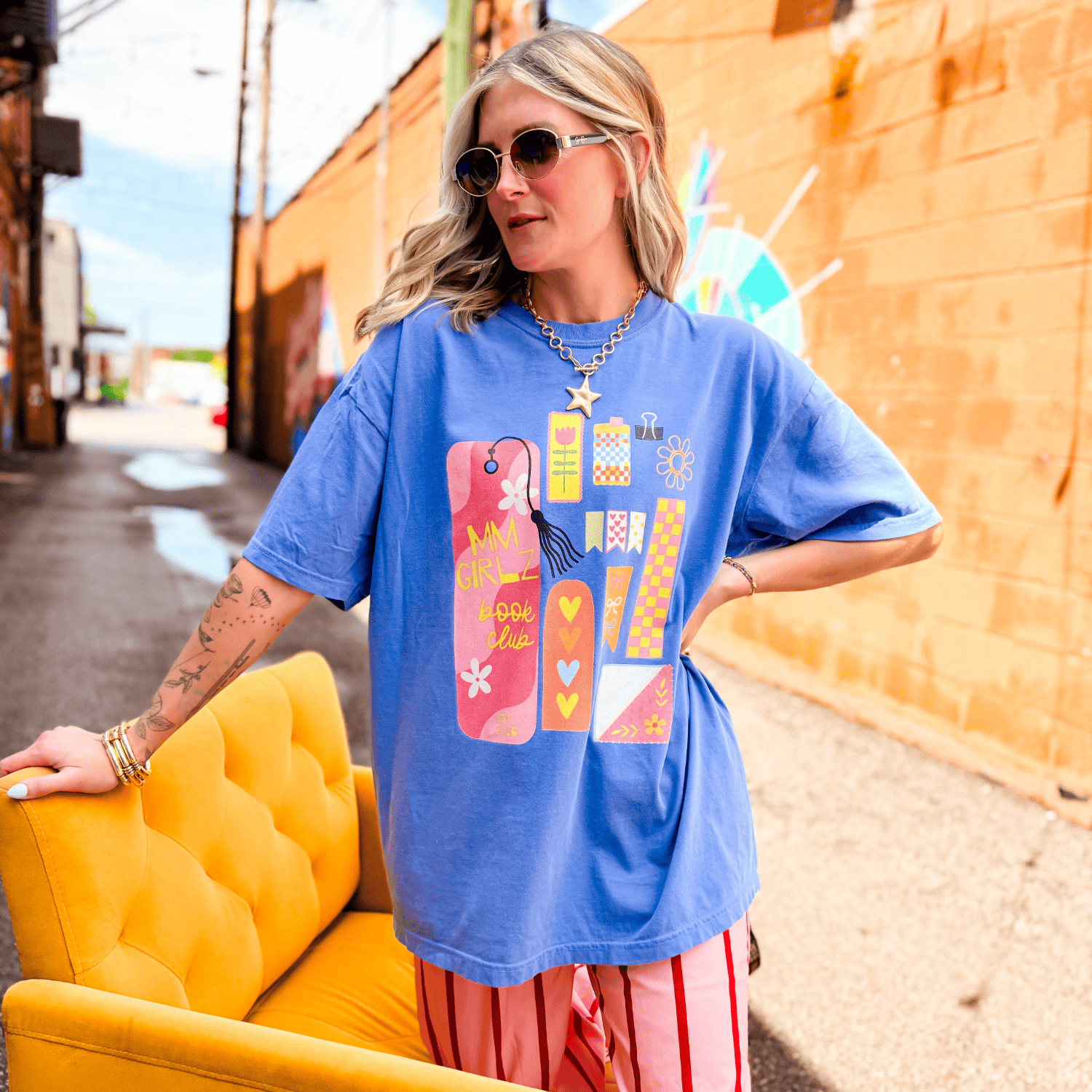 Blue Book Club Tee featuring colorful graphic designs, worn by a woman seated on a yellow chair in an urban setting.