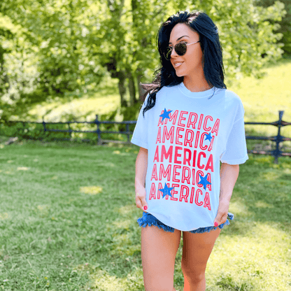 Starry America tee in white with red and blue text, worn by a woman outdoors with sunglasses and denim shorts.