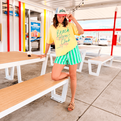 Yellow Beach Mom's Club tee paired with green striped shorts, worn by a woman posing on a wooden bench.