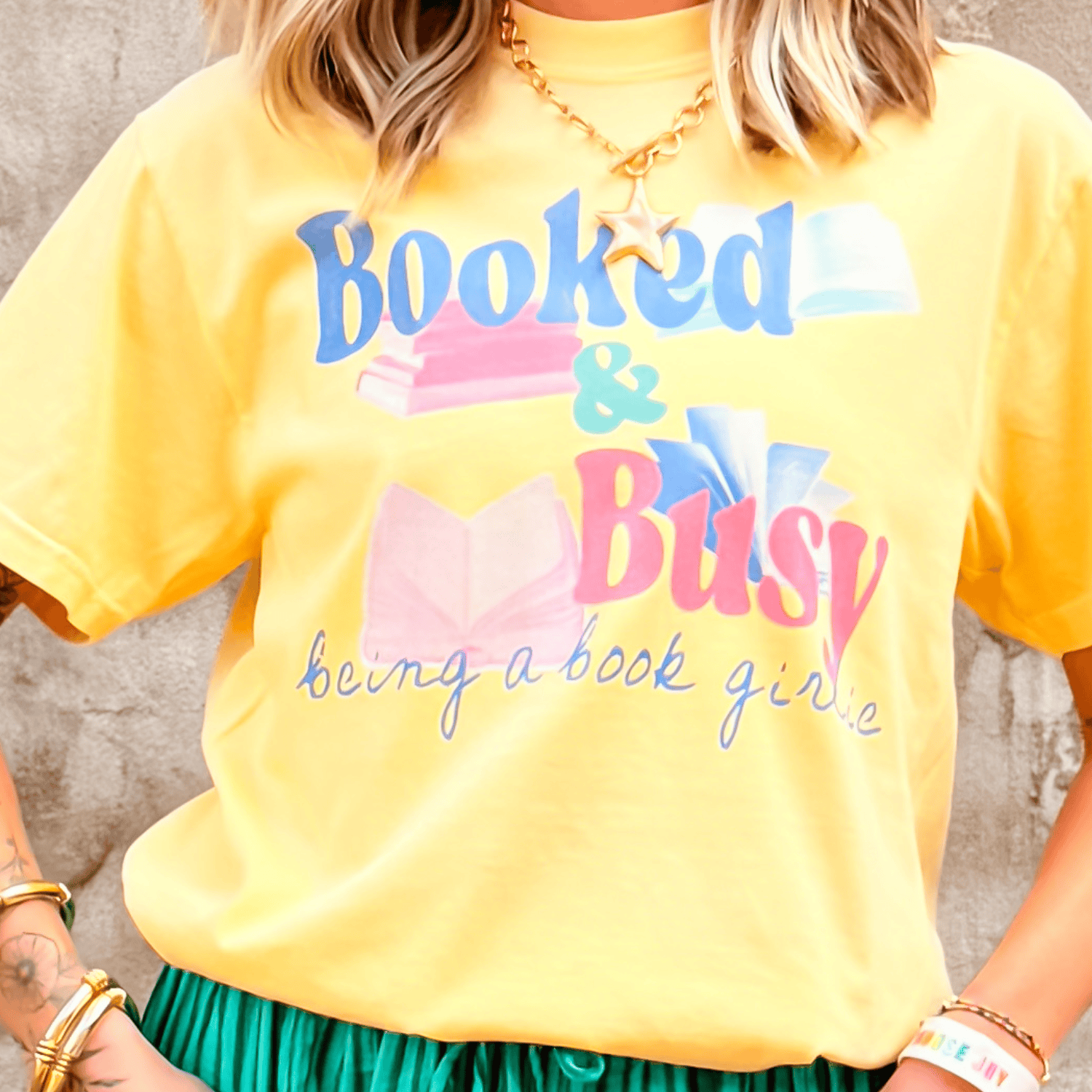 Booked & Busy tee in yellow featuring colorful book graphics and text, styled with a green skirt and accessories.
