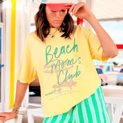 Yellow Beach Mom's Club tee with green text, worn by a model with a pink cap, paired with striped green shorts.