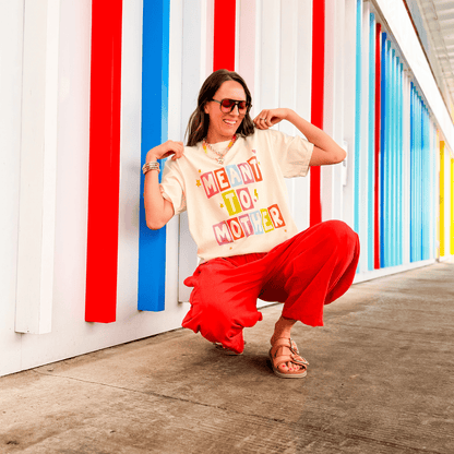 Meant To Mother tee in cream with colorful lettering, worn by a woman squatting against a brightly colored wall.