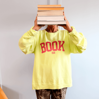 Crewneck sweatshirt in neon yellow with "BOOKISH" printed in red, worn by a person holding stacked books.