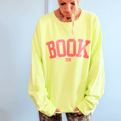 Bright neon yellow crewneck sweatshirt featuring bold pink "BOOK" and "ISH" text, worn by a person with long hair.