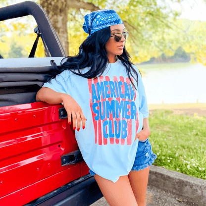 Light blue American Summer Club tee with bold red lettering, worn by a woman beside a red vehicle in a sunny outdoor setting.